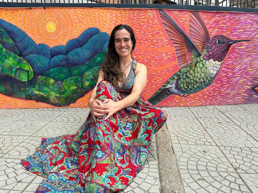 Cata Heincke sentada frente a un mural colorido con un colibrí, representando su servicio al renacimiento, la transformación y la conexión con el alma.